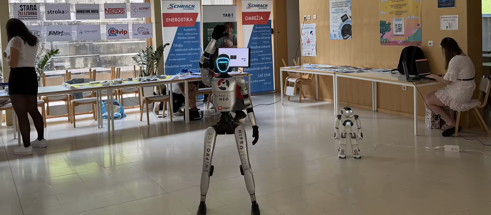 Robot NEO stoji med skupino dijakov na šolskem hodniku.
