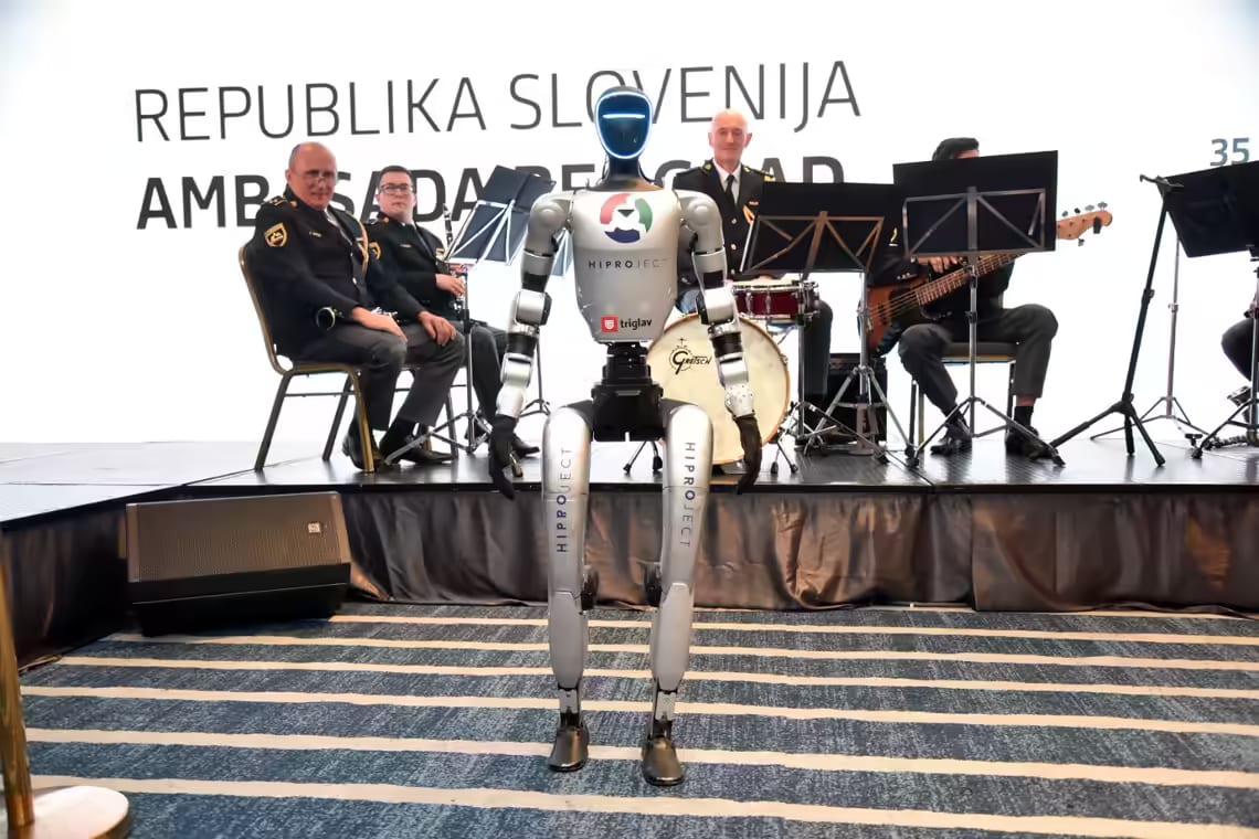 Humanoidni robot NEO stoji pred pihalnim orkestrom na sprejemu z napisom Republika Slovenija