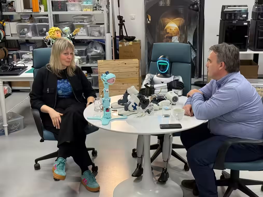 Humanoidni robot sedi za mizo z dvema obiskovalcema v delavnici s 3D tiskalniki