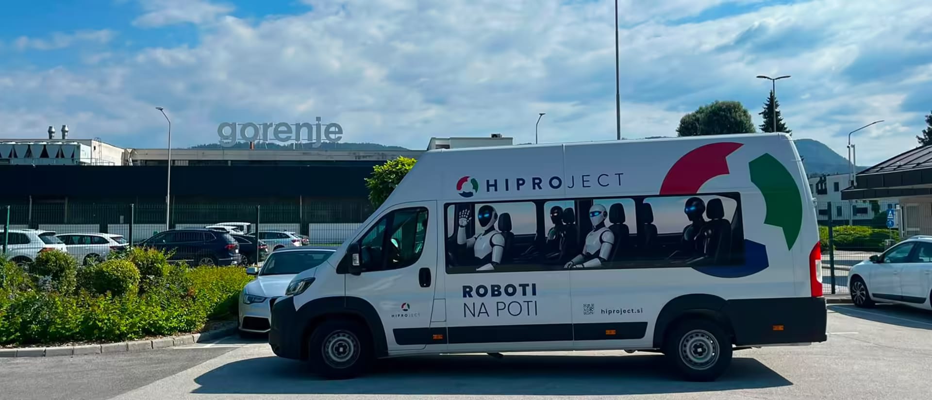 Kombi HIPROJECT z napisom ROBOTI NA POTI in humanoidnimi roboti na sedežih, parkiran pred industrijskim obratom