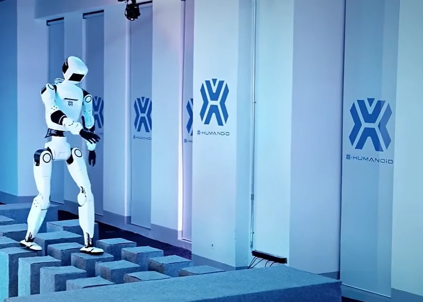 Robot OMNI stopa po razmaknjenih kockah različnih višin v prostoru z logotipi X-HUMANOID na stenah