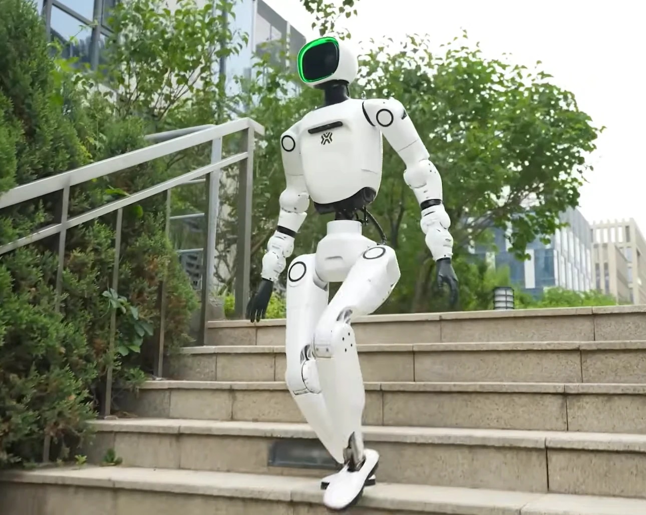 Robot OMNI hodi po zunanjih kamnitih stopnicah med drevesi, glava z zelenim obročem obrnjena v smer hoje