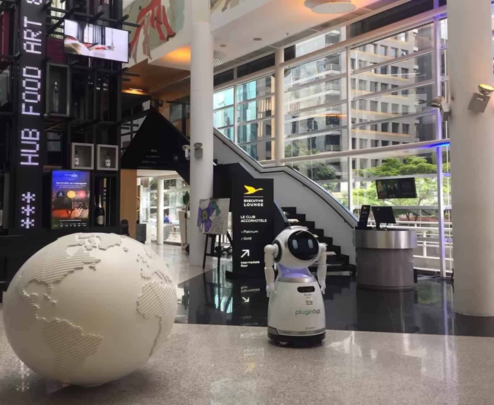 Servisni robot na recepciji hotelske avle, ob njem globus in stopnišče.